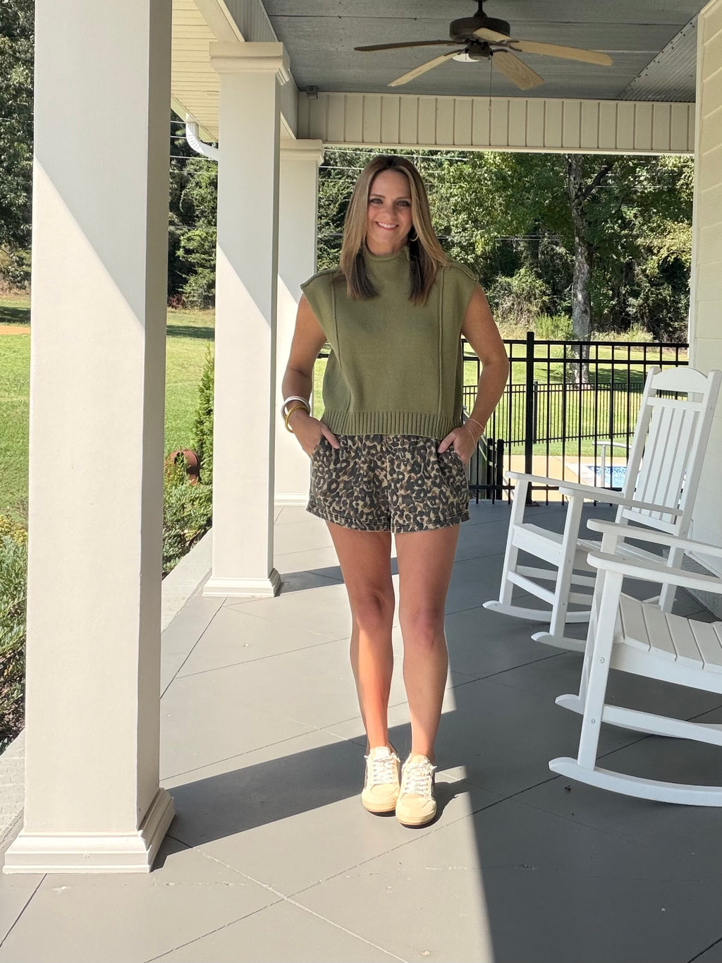 She’s wearing an olive green sleeveless mock-neck knit sweater top and leopard-print shorts, paired with white sneakers. 